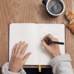 Open journal on table, hands on top, right hand writing, one croissant next to journal with a bite taken out, and a cup of coffee nearby