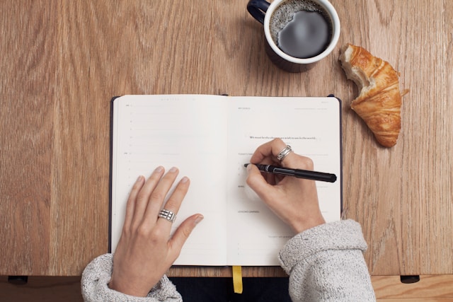 Open journal on table, hands on top, right hand writing, one croissant next to journal with a bite taken out, and a cup of coffee nearby